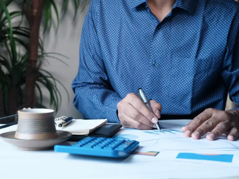 Um homem em camisa social azul examinando documentos e gráficos com uma caneta, com uma calculadora e xícara de café na mesa